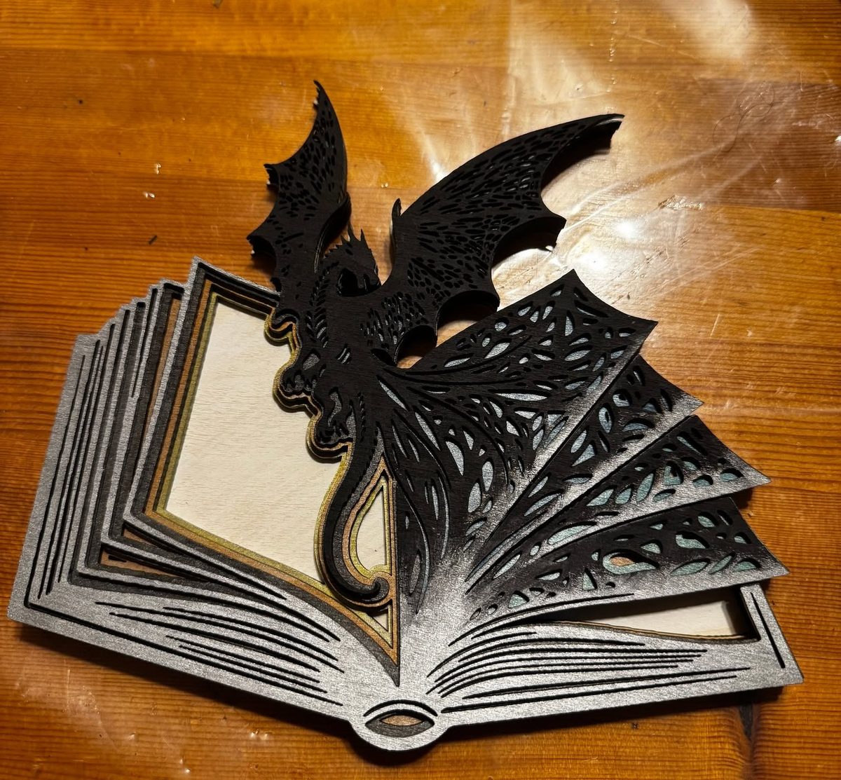 A layered piece shaped like an open book with its pages fanned, a winged dragon perched on the spine, cut in black and white and standing on a wooden table.