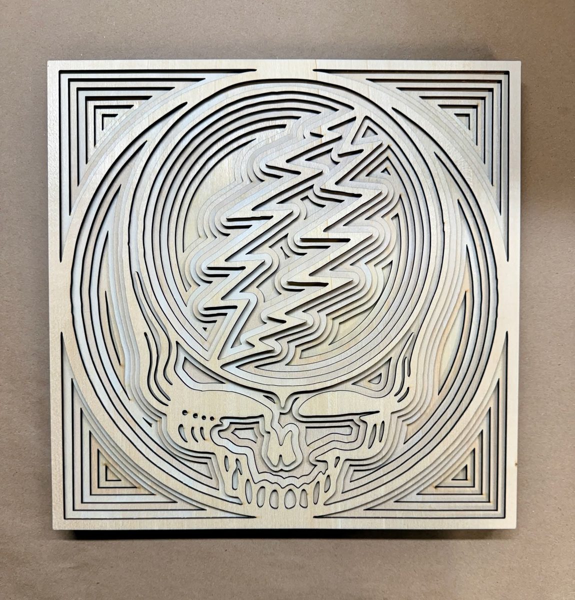 A square layered panel in pale wood: a skull split down the middle by a lightning bolt, inside a stepped square border.