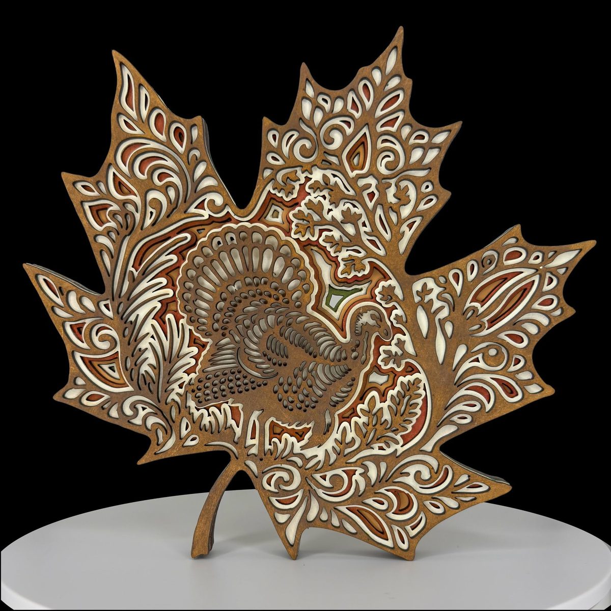 A maple-leaf-shaped layered piece in natural wood on a black background, standing on a small easel, with a turkey among ferns at its centre.