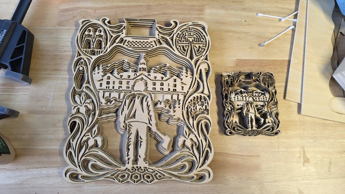 Two versions of the same layered panel on a workbench, one about twice the size of the other. A figure walks away from the viewer towards a long mountain lodge with rows of lit windows, pine trees and peaks behind it. Art-nouveau stems and irises frame the scene, and four small medallions around the edge hold two figures side by side, a typewriter, a hedge maze and an axe.