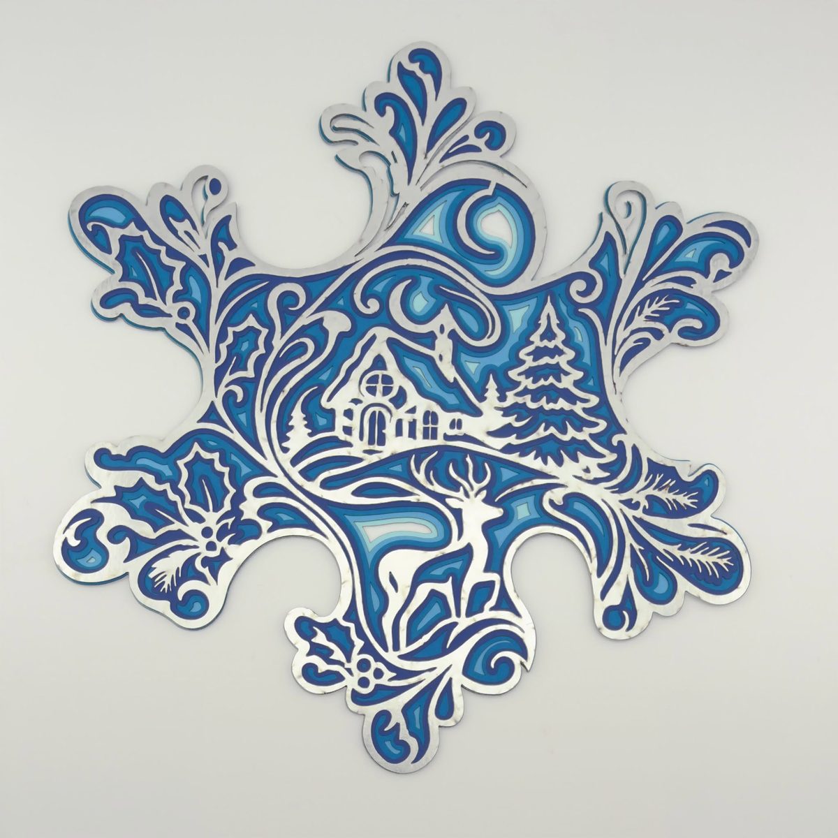 A snowflake-shaped layered piece in blues and white, holding a winter village scene with a church, fir trees and a deer.