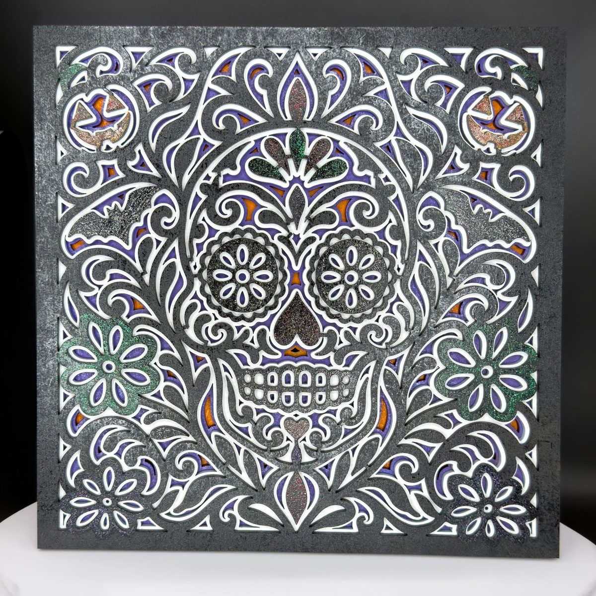 A square layered panel with a black frame and white and silver interior layers, showing a sugar skull surrounded by scrollwork and small flowers.